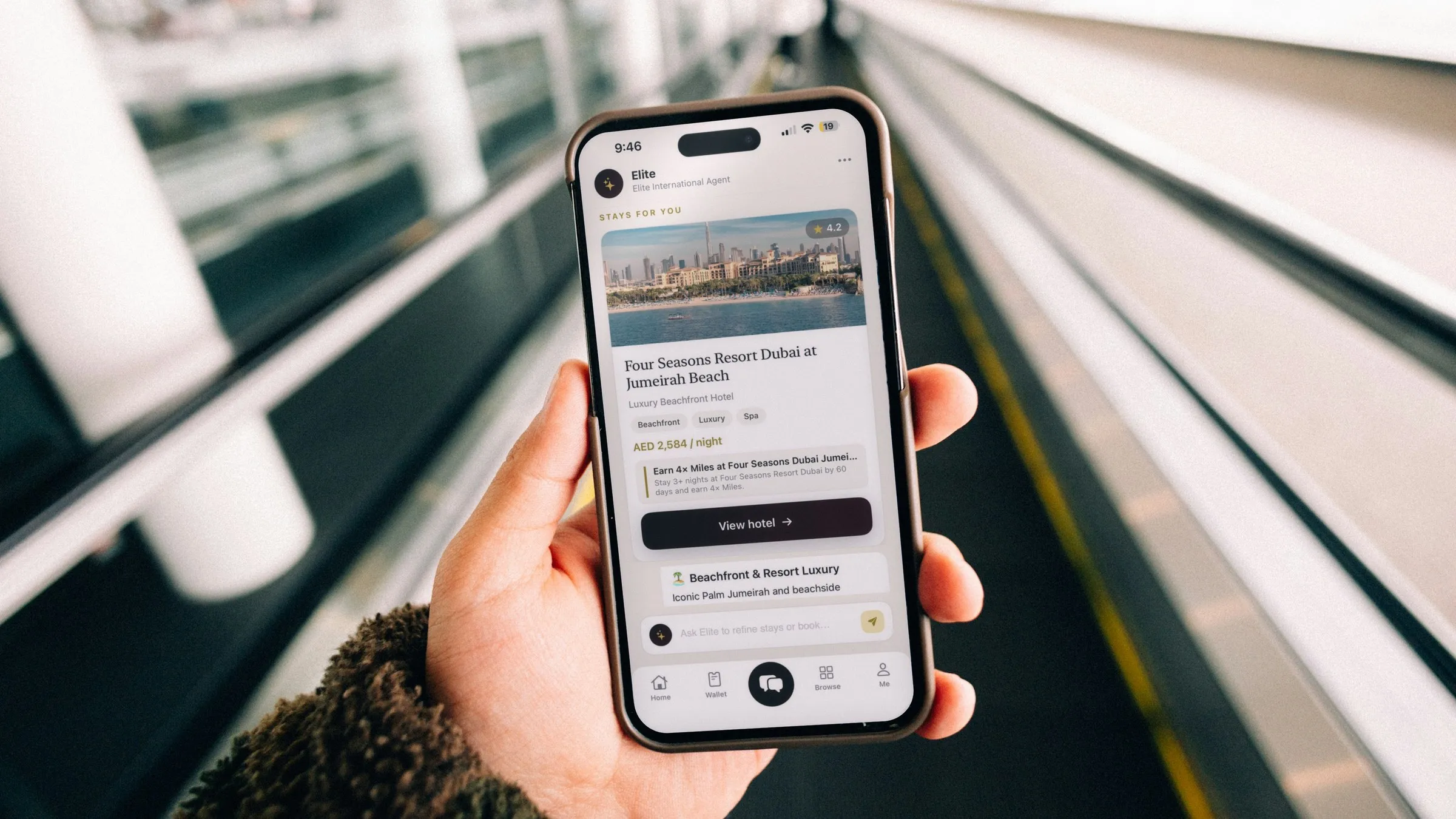 A hand holds a smartphone in an airport walkway lined with moving walkways. On the screen, the Elite agent suggests the Four Seasons Resort Dubai at Jumeirah Beach, with an offer to earn 4x Miles on a stay of three nights or more.