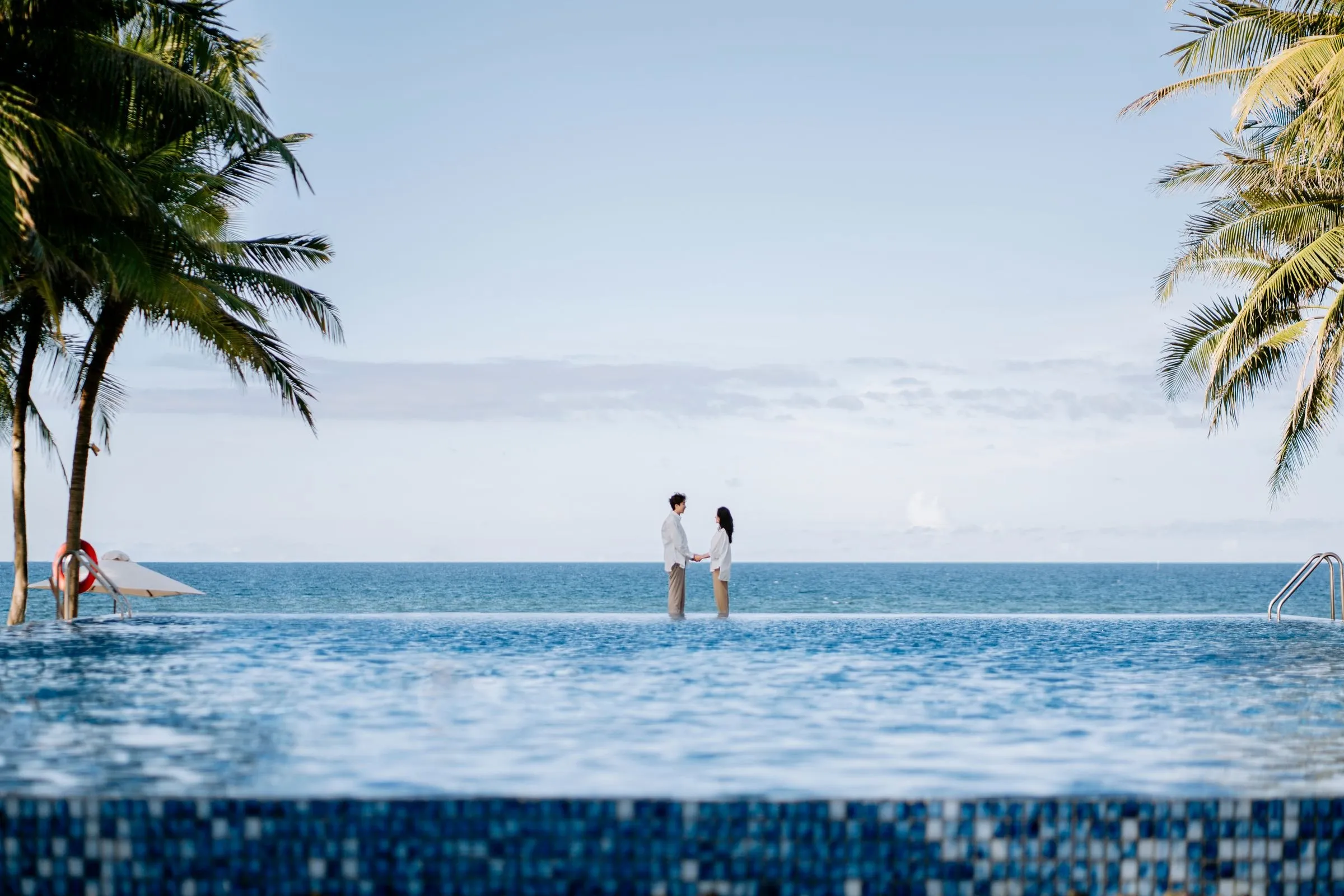 A couple stands at the edge of an infinity pool that meets the ocean, framed by palm trees.