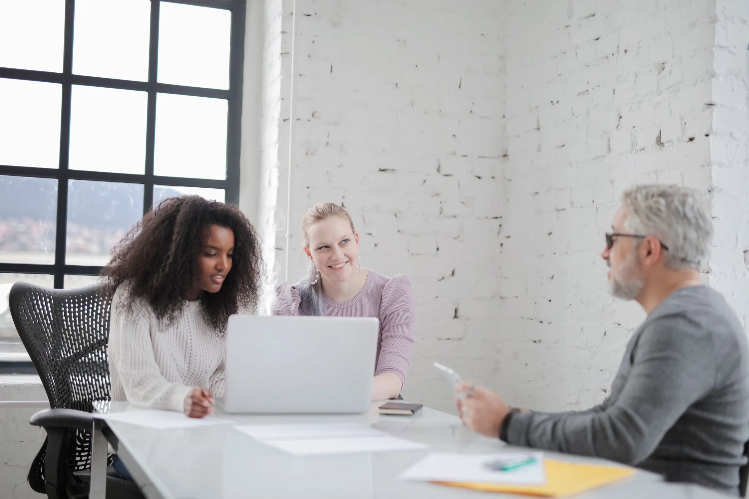 Three colleagues review a laptop together at a white table in a bright brick-walled office.