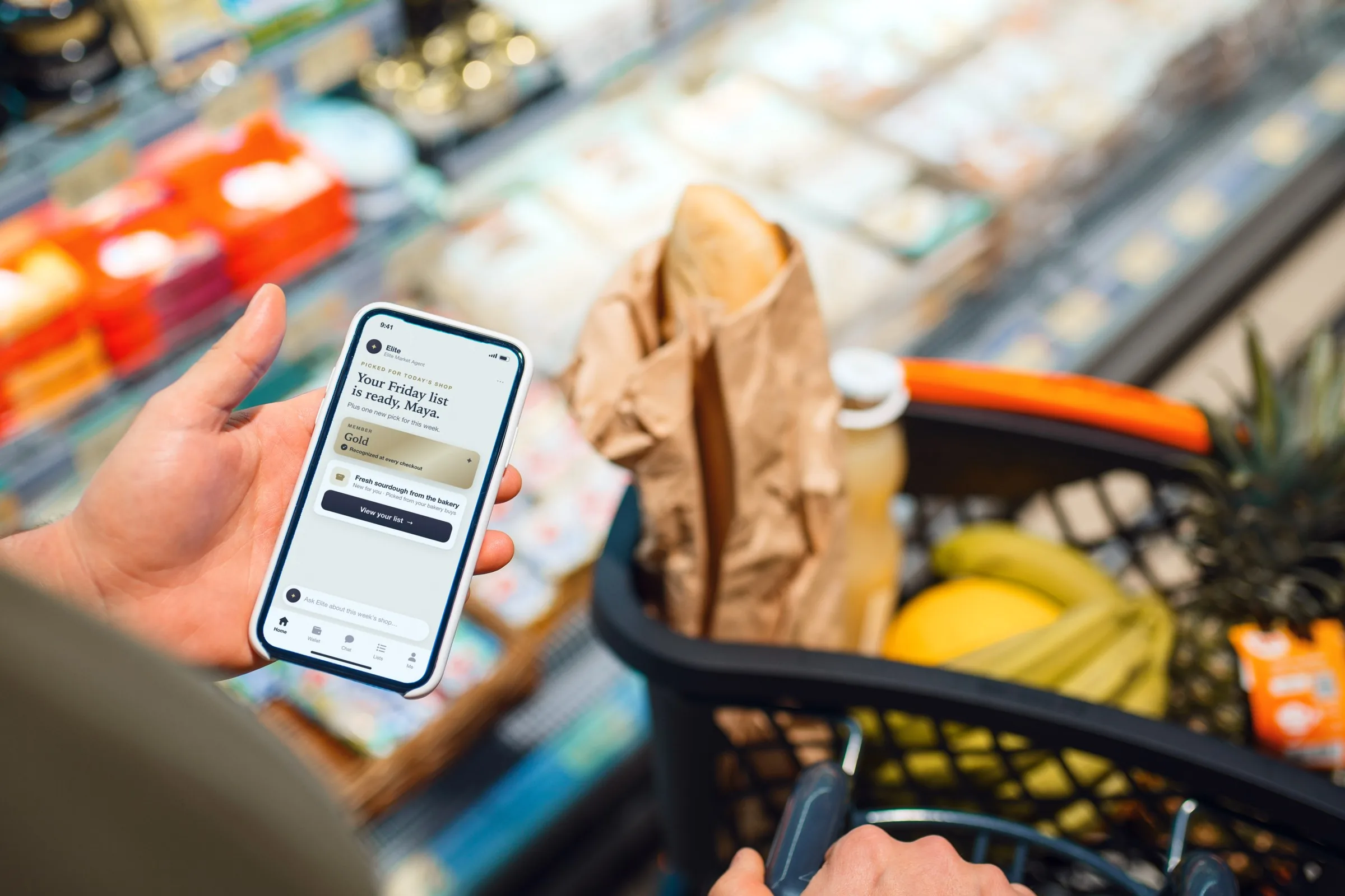 A shopper holds a phone above a grocery cart with a bakery loaf and fruit. The app says the Friday list is ready, recognizes Gold status and adds one new pick: fresh sourdough from the bakery.