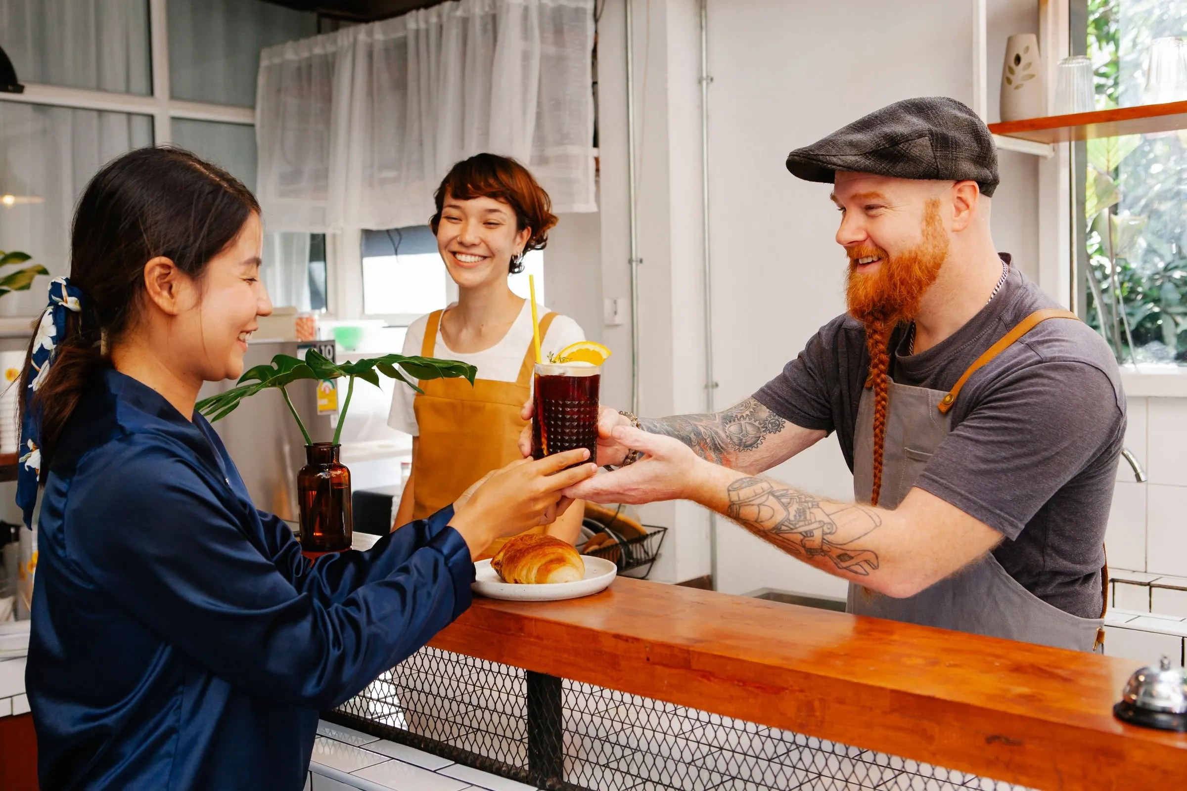 A smiling barista hands a regular her iced drink across a cafe counter while a colleague looks on.