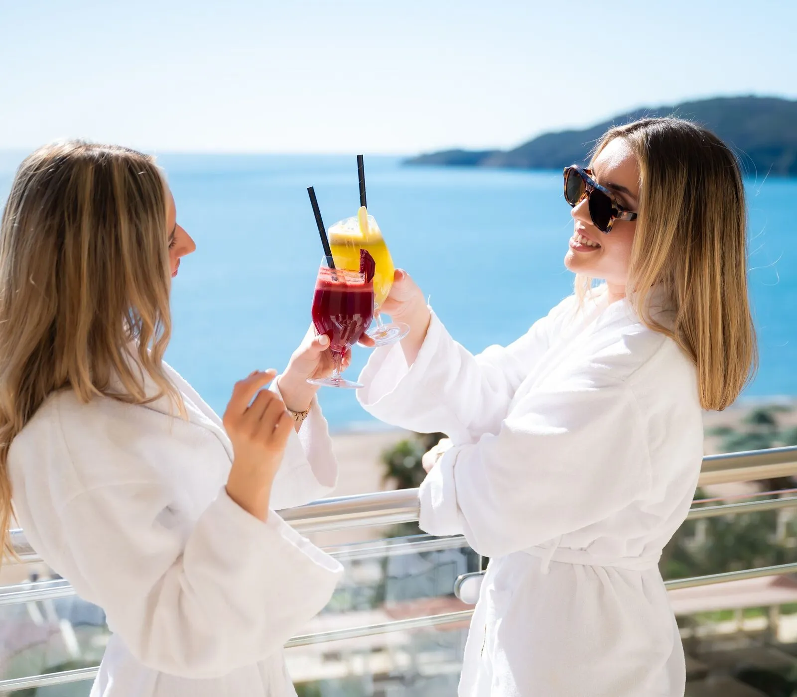 Two friends in white bathrobes toast with drinks on a sunny balcony above the sea.