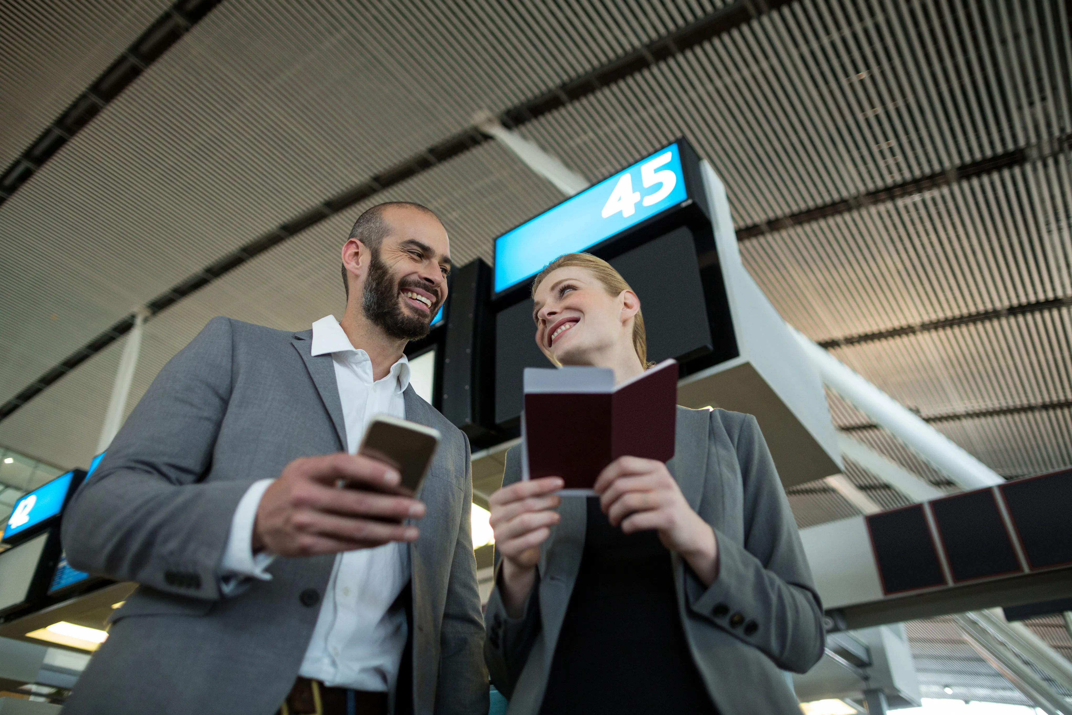Two travelers smiling at an airport gate, one holding a phone and the other a passport