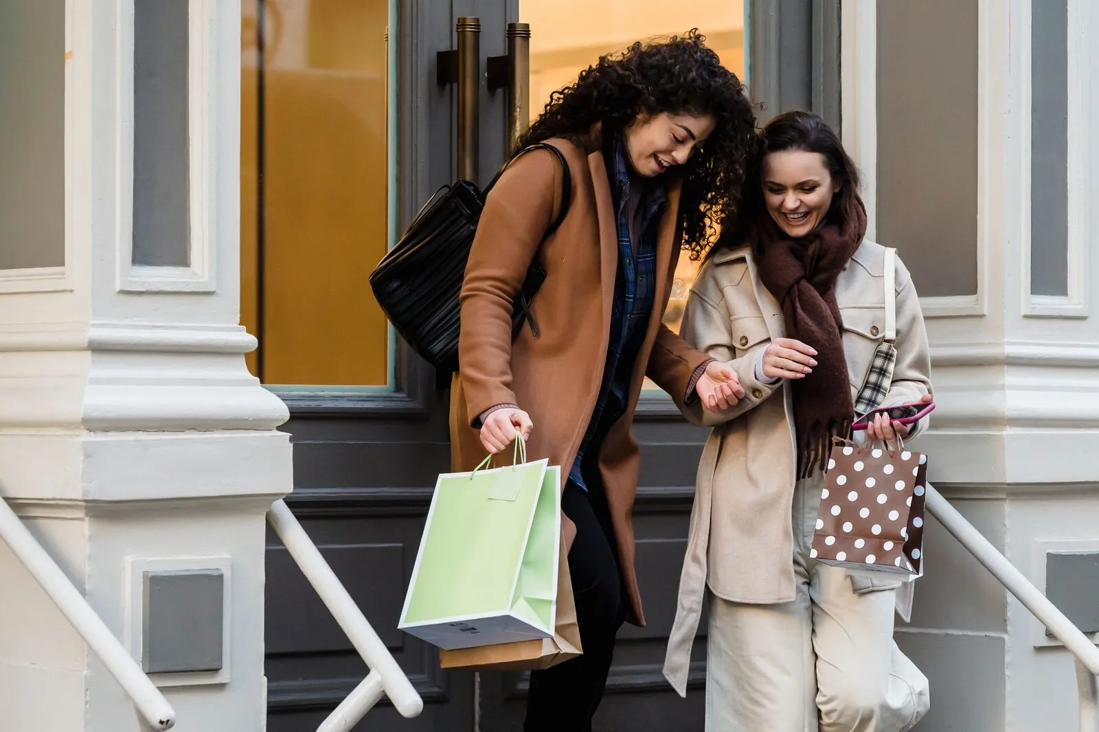 Two friends laugh as they step out of a store carrying shopping bags.